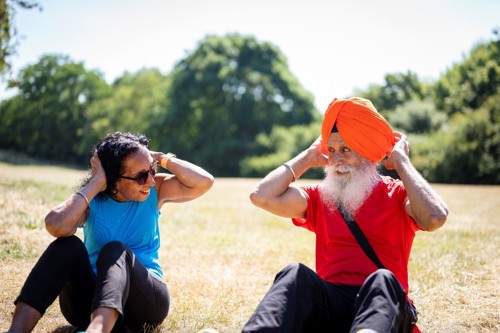 An Asian man and women in their 60s or 70s are exercising outside on a warm, sunny day. They are wearing colourful exercise clothes and smiling at each other. They are enjoying themselves.